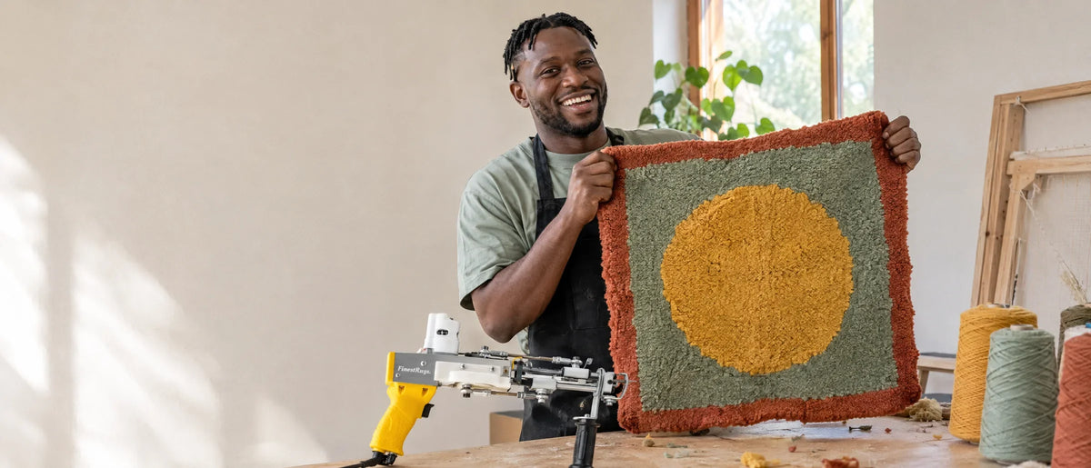 Tufter houdt lachend zijn eerste, iets scheve zelfgemaakte kleed omhoog, tufting gun op tafel